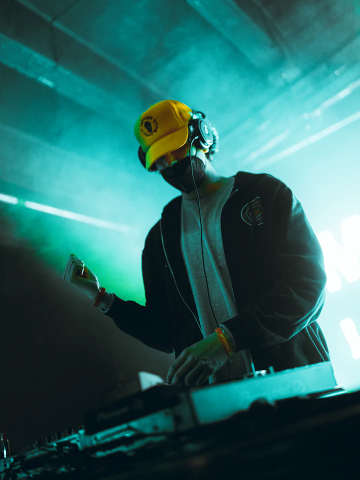 A DJ in a yellow cap and headphones mixing under green club lights